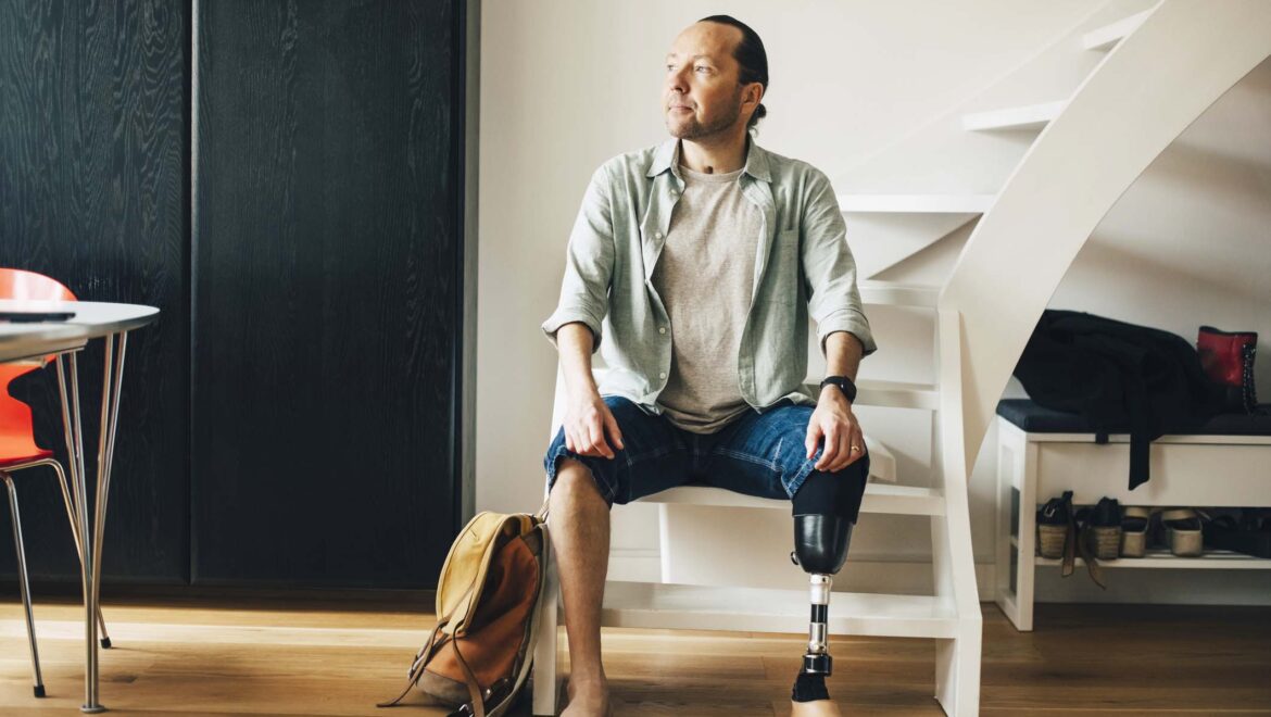 Man with a prosthetic leg sitting on stairs inside his apartment and looking away from the camera.